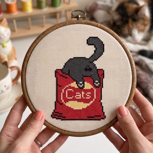 K&ouml;nnte beinhalten: Ein runder Holzstickrahmen mit einem Kreuzstichmotiv, das eine schwarze Katze zeigt, die aus einer roten T&uuml;te mit einem gelben kreisf&ouml;rmigen Emblem herausragt. Der Text "Cats" ist in wei&szlig;en Buchstaben im gelben Kreis auf der T&uuml;te gestickt.