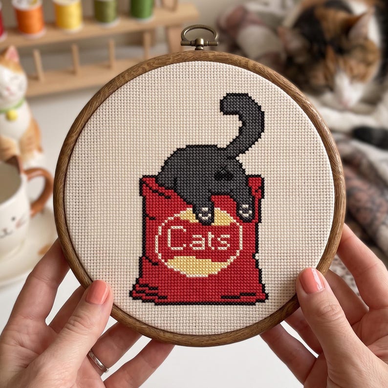 K&ouml;nnte beinhalten: Ein runder Holzstickrahmen mit einem Kreuzstichmotiv, das eine schwarze Katze zeigt, die aus einer roten T&uuml;te mit einem gelben kreisf&ouml;rmigen Emblem herausragt. Der Text "Cats" ist in wei&szlig;en Buchstaben im gelben Kreis auf der T&uuml;te gestickt.