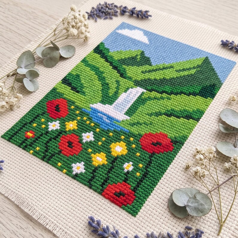 Pu&ograve; includere: Un ricamo a punto croce su tessuto color crema raffigurante un paesaggio con colline verdi, una cascata bianca, un cielo azzurro con una nuvola bianca e un primo piano pieno di papaveri rossi, fiori gialli e margherite bianche.