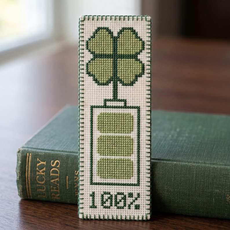 May include: A rectangular bookmark featuring a cross-stitch design on cream fabric. The design displays a four-leaf clover above a battery icon with three filled segments, and the text 100% at the bottom, all stitched in shades of green.
