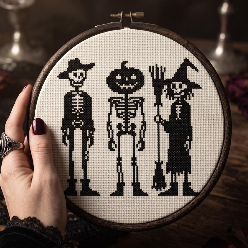 K&ouml;nnte beinhalten: Ein runder Holzstickrahmen mit einem Kreuzstichmuster auf wei&szlig;em Stoff. Das Design zeigt drei schwarze Figuren: ein Skelett mit Hut, ein Skelett mit K&uuml;rbiskopf und eine Hexe, die einen Besen h&auml;lt.