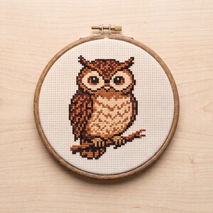 May include: A circular wooden embroidery hoop containing a cross-stitch pattern of a brown and tan owl perched on a branch. The owl features large, round eyes and detailed feather textures, centered on a cream-colored fabric background.