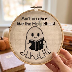 May include: A circular wooden embroidery hoop containing a piece of cream-coloured Aida fabric with black cross-stitch embroidery. The design features a cartoon ghost holding a black book with a cross, and the text: Ain't no ghost like the Holy Ghost.