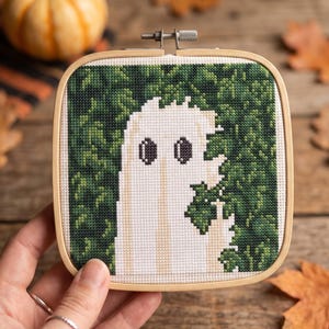 May include: A square wooden embroidery hoop containing a cross-stitch pattern of a white ghost figure with dark oval eyes, positioned against a dense background of dark green foliage. The ghost is depicted with a raised hand showing a middle finger.