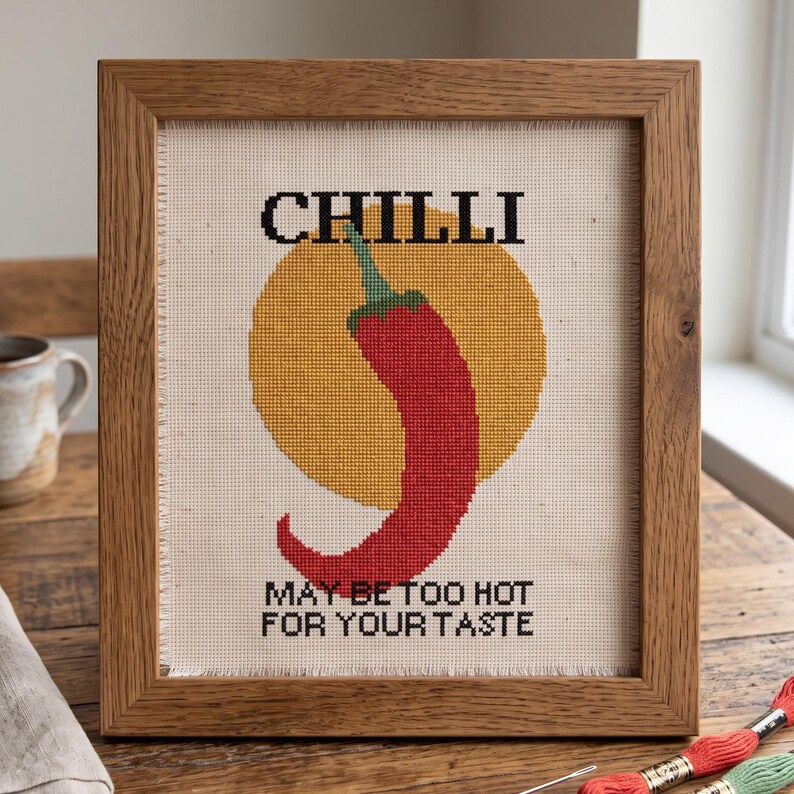 Pu&ograve; includere: Un ricamo a punto croce incorniciato raffigurante un peperoncino rosso centrato su un cerchio giallo su tessuto color crema. Il testo CHILLI &egrave; ricamato in nero sopra il peperoncino e il testo MAY BE TOO HOT FOR YOUR TASTE &egrave; ricamato in nero sotto.