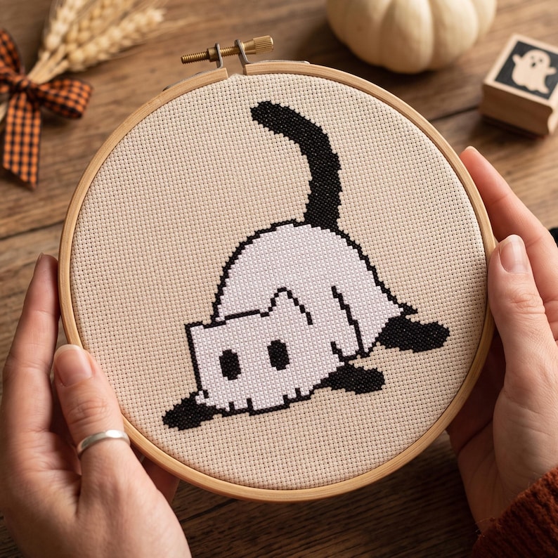May include: A circular wooden embroidery hoop containing a piece of beige Aida fabric with a cross-stitch design of a white cat with black ears, a black tail, and black paws, depicted in a playful pouncing position on the fabric surface.
