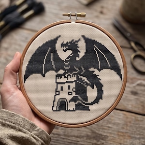 Peut inclure: Un tambour &agrave; broder circulaire en bois contenant un motif au point de croix sur tissu beige. Le design pr&eacute;sente la silhouette noire d'un dragon perch&eacute; au sommet d'une tour en pierre. Le dragon a de grandes ailes d&eacute;ploy&eacute;es et une longue queue.