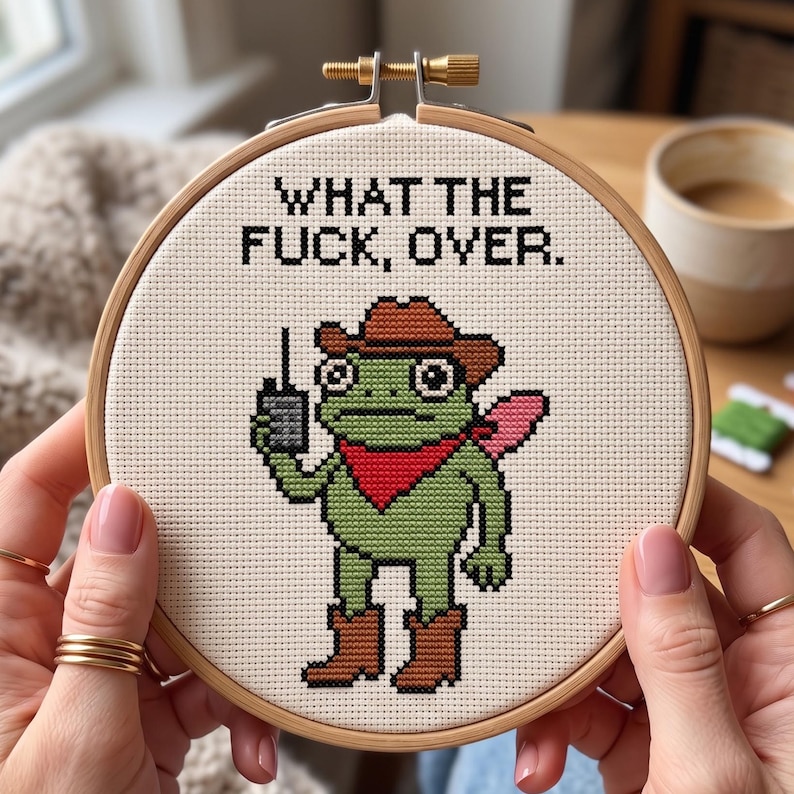 May include: A circular wooden embroidery hoop containing a cross-stitch design on off-white fabric. The design features a green frog wearing a brown cowboy hat, brown boots, and a red bandana, holding a walkie-talkie. The text reads: WHAT THE FUCK, OVER.
