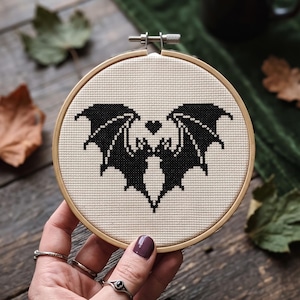 Puede incluir: Un bastidor de bordado circular de madera que contiene un dise&ntilde;o de punto de cruz sobre tela de color crema. El dise&ntilde;o presenta la silueta sim&eacute;trica de un murci&eacute;lago negro con las alas extendidas y un peque&ntilde;o coraz&oacute;n negro entre las orejas.