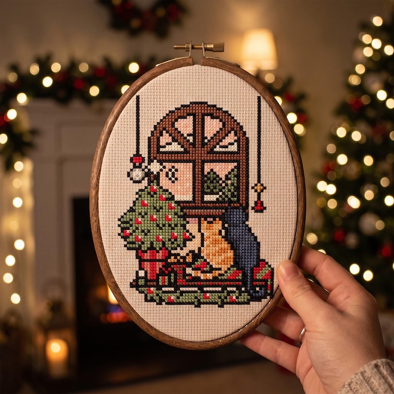 K&ouml;nnte beinhalten: Ein ovaler Stickrahmen aus Holz mit einem Kreuzstichmuster, das ein Fenster, einen geschm&uuml;ckten Weihnachtsbaum und zwei zusammensitzende Katzen zeigt. Das Design umfasst einen braunen Fensterrahmen, einen gr&uuml;nen Baum und orangefarbene und blaue Katzen.
