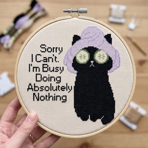 May include: A circular wooden embroidery hoop containing a cross-stitch design on cream fabric. The design features a black cat wearing a light purple towel wrap on its head and green cucumber slices over its eyes, with the text Sorry I Can't. I'm Busy Doing Absolutely Nothing in black.