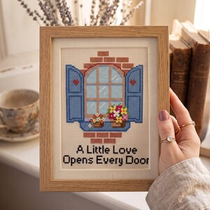 Peut inclure: Une broderie au point de croix encadr&eacute;e repr&eacute;sentant un mur de briques avec une fen&ecirc;tre ouverte, des volets bleus et des jardini&egrave;res. Le motif inclut des c&oelig;urs rouges et le texte A Little Love Opens Every Door en noir sur fond clair.