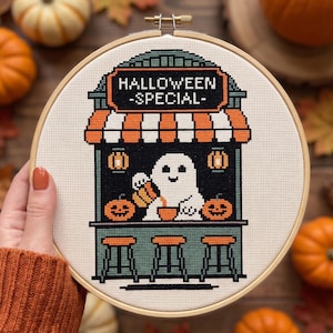 May include: A circular wooden embroidery hoop containing a cross-stitch design of a ghost behind a counter. The text reads HALLOWEEN SPECIAL. The scene features orange and white striped awnings, two jack-o'-lanterns, and three bar stools.