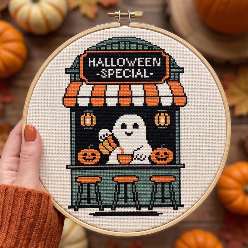 May include: A circular wooden embroidery hoop containing a cross-stitch design of a ghost behind a counter. The text reads HALLOWEEN SPECIAL. The scene features orange and white striped awnings, two jack-o'-lanterns, and three bar stools.