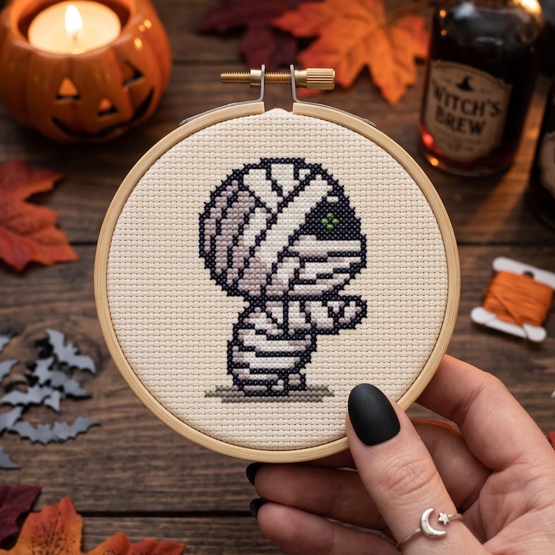 May include: A circular wooden embroidery hoop containing a cross-stitch design of a cartoon mummy. The mummy is stitched in shades of light gray and white with a single green eye, set against a cream-colored fabric background with a small shadow.