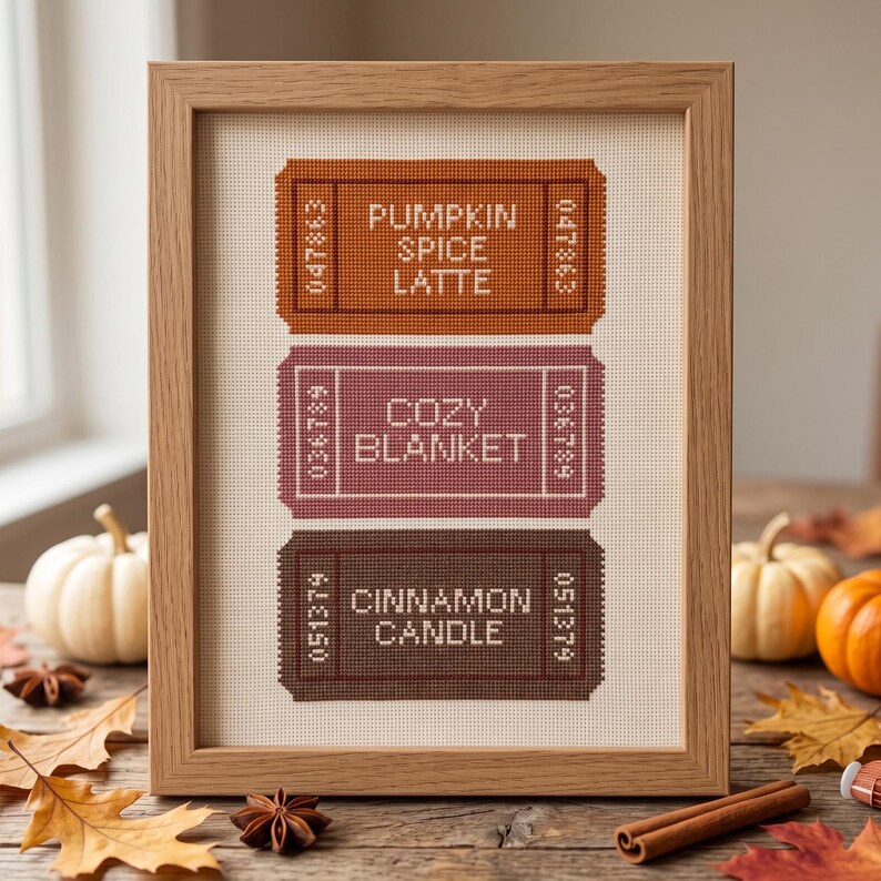 May include: A framed cross-stitch embroidery featuring three rectangular ticket-shaped designs in orange, mauve, and dark brown. The text reads PUMPKIN SPICE LATTE, COZY BLANKET, and CINNAMON CANDLE, each accompanied by unique numerical codes on the sides.