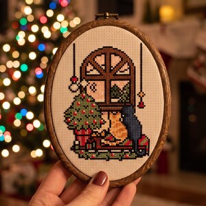 K&ouml;nnte beinhalten: Ein ovaler Stickrahmen aus Holz mit einem Kreuzstichmuster, das ein Fenster, einen geschm&uuml;ckten Weihnachtsbaum und zwei Katzen, eine orangefarbene und eine dunkelblaue, auf einem Sims sitzend zeigt. Der Stoff ist cremefarben und kariert.