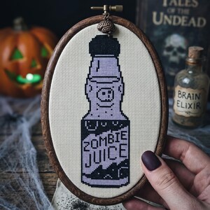 K&ouml;nnte beinhalten: Ein ovaler Stickrahmen aus Holz mit einem Kreuzstichmotiv einer Flasche, die mit ZOMBIE JUICE in schwarzem und hellviolettem Garn beschriftet ist. Ein kleiner gehirnf&ouml;rmiger Anh&auml;nger ist oben am Rahmen des Stickrahmens befestigt.