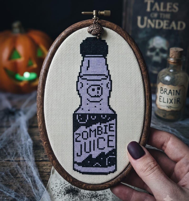 K&ouml;nnte beinhalten: Ein ovaler Stickrahmen aus Holz mit einem Kreuzstichmotiv einer Flasche, die mit ZOMBIE JUICE in schwarzem und hellviolettem Garn beschriftet ist. Ein kleiner gehirnf&ouml;rmiger Anh&auml;nger ist oben am Rahmen des Stickrahmens befestigt.