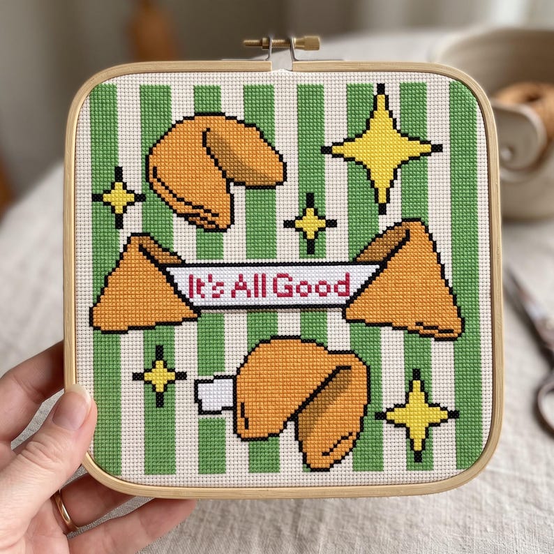 May include: A square wooden embroidery hoop containing cross-stitch fabric with a green and white vertical striped pattern. The design features orange fortune cookies, yellow four-pointed stars, and a white banner with the text It's All Good.