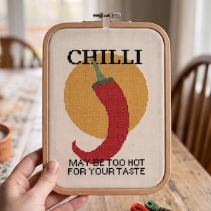 Pu&ograve; includere: Un telaio da ricamo rettangolare in legno contenente un disegno a punto croce su tessuto bianco. Il motivo raffigura un peperoncino rosso su un cerchio giallo, con il testo nero CHILLI e MAY BE TOO HOT FOR YOUR TASTE ricamato sopra e sotto.