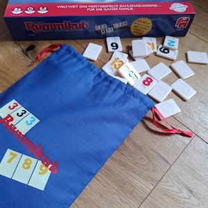 Op de afbeelding: Een Rummikub bordspelset met een blauwe zak met trekkoord, tegels en een doos. De zak toont de naam van het spel en tegelcombinaties. De tegels zijn wit met gekleurde cijfers. De doos is rood en blauw.