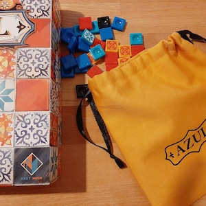 Könnte beinhalten: Eine gelbe Kordeltasche mit dem Wort "Azul" darauf gedruckt. Die Tasche liegt neben einer Schachtel des Brettspiels Azul und einem Stapel bunter Spielsteine.