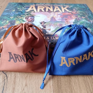 Puede incluir: Dos bolsas con cordón, una de color óxido y otra azul, con la palabra "Arnak" impresa. Las bolsas están delante de una caja de juego de mesa con el título "Arnak" e ilustraciones de exploradores.