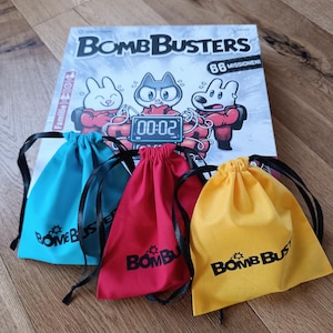 Peut inclure: Trois sacs à cordon de couleur avec le logo "Bomb Busters" imprimé dessus. Les sacs sont devant une boîte de jeu de société avec le titre "Bomb Busters" et le texte "88 Missionen!" imprimé dessus.