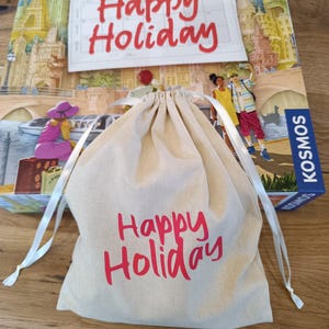 Könnte beinhalten: Ein Brettspielkarton mit einer farbenfrohen Illustration berühmter Sehenswürdigkeiten und den roten Worten "Happy Holiday". Vor der Schachtel befindet sich ein beigefarbener Kordelzugbeutel mit demselben Text. Die Spielbox hat die Marke "KOSMOS".