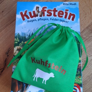 Könnte beinhalten: Eine grüne Kordeltasche mit dem Text "Kuhfstein" in braunen Buchstaben und einer weißen Kuh-Silhouette. Die Tasche liegt auf einer Brettspielbox mit dem Titel "Kuhfstein" in roten Buchstaben und dem Text "Hegen, pflegen, Felder legen!" in schwarzen Buchstaben.