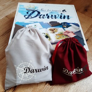 Peut inclure: Deux sacs à cordon, l'un beige et l'autre bordeaux, avec le mot "Darwin" imprimé dessus. Le sac beige est orné d'une boussole et le sac bordeaux d'une police d'écriture cursive simple. Les sacs sont posés sur une surface en bois devant une boîte de jeu de société intitulée "Sur les traces de Darwin".
