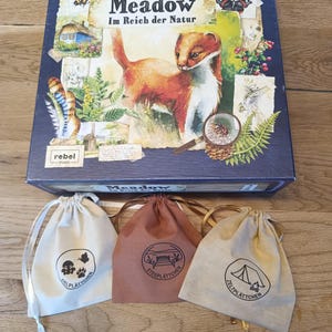 Pode incluir: Uma caixa de jogo de tabuleiro quadrada intitulada Meadow Im Reich der Natur com uma ilustração em aguarela de uma doninha, insetos e plantas, acompanhada por três pequenos sacos de tecido com cordão em creme, castanho e bege com ícones.