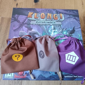 Op de afbeelding: Drie stoffen zakken met verschillende symbolen erop. De zakken zijn bruin, grijs en paars. De zakken liggen voor een bordspeldoos met de titel "Klong! Katakomben".