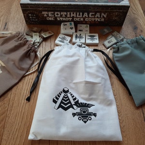 Puede incluir: Tres bolsas de cordón con diferentes diseños. La bolsa blanca tiene una pirámide negra y una figura estilizada negra. La bolsa marrón tiene una figura estilizada dorada. La bolsa azul tiene una figura estilizada dorada.