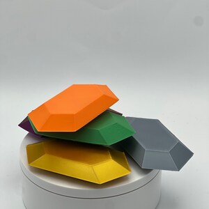 Pu&ograve; includere: Una pila di cinque oggetti stampati in 3D di forme geometriche e colori vivaci. Gli oggetti sono disposti a forma di piramide, con l'oggetto pi&ugrave; grande in basso e quello pi&ugrave; piccolo in alto. Gli oggetti sono realizzati in un materiale liscio, simile alla plastica. I colori degli oggetti sono arancione, verde, grigio, oro e viola.