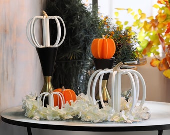 May include: Three white pumpkin-shaped decorations on a table with a white floral arrangement. The pumpkins are made of wood and have a geometric design. The center pumpkin is on a black stand with a gold base.