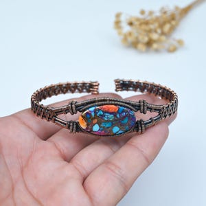 Könnte beinhalten: Ein Armreif aus dunkel kupferfarbenem Draht mit einem zentralen, ovalen Stein, der ein lebhaftes Mosaikmuster in Türkis, Orange, Violett und Braun aufweist und von kunstvollen, gewickelten Metalldrähten gehalten wird.