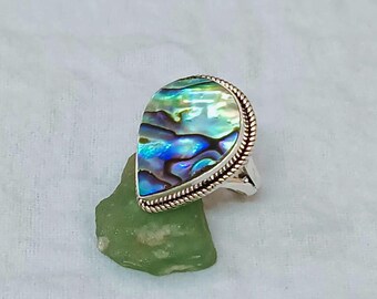 Könnte beinhalten: Ein silberner Ring mit einer tropfenförmigen Abalone-Muschel. Die Muschel zeigt schillernde Blau-, Grün- und Lilatöne, eingefasst von einem gedrehten Seilmuster. Der Ring liegt auf einem grünen Stein vor weißem Hintergrund.