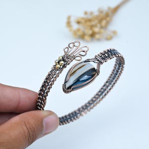 May include: A wire-wrapped cuff bracelet crafted from copper wire, featuring a central teardrop-shaped banded agate stone with grey and white layers. The design includes intricate woven wire patterns, decorative loops, and small brass-colored metal beads.