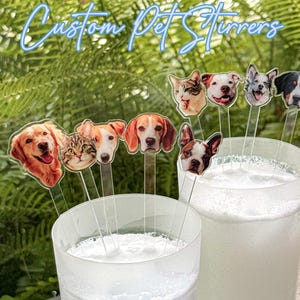 May include: Clear acrylic drink stirrers featuring custom printed pet faces, including dogs and cats, with the text Custom Pet Stirrers displayed at the top. The stirrers are placed inside two clear glasses filled with a white, frothy liquid beverage.
