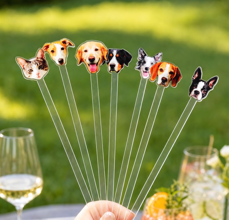 May include: A set of seven clear acrylic drink stirrers, each topped with a different animal face including a cat and various dog breeds such as a golden retriever, a beagle, and a Boston terrier, arranged in a fan shape against a blurred background.