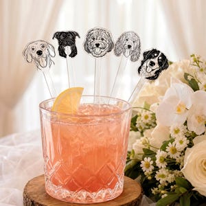 May include: Five clear acrylic drink stirrers featuring black and white line art illustrations of different dog breeds, placed inside a textured glass filled with a pink beverage, ice cubes, and a single yellow lemon slice wedge.