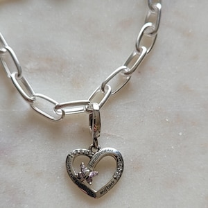Könnte beinhalten: Ein silberfarbenes Charm-Armband mit einem herzförmigen Anhänger. Der Anhänger trägt die Aufschrift "I love you to the moon and back Granddaughter" und zeigt eine kleine rosa Blume und klare Kristalle.