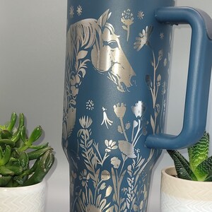 May include: A blue stainless steel travel mug with a silver horse and floral design. The mug has a lid and a handle.