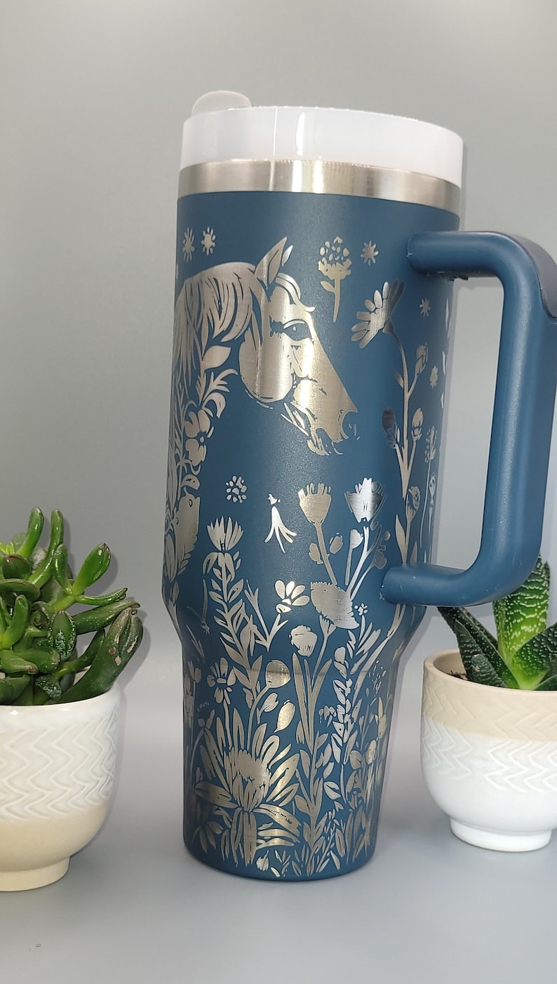 May include: A blue stainless steel travel mug with a silver horse and floral design. The mug has a lid and a handle.
