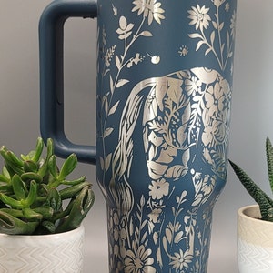 May include: A blue stainless steel travel mug with a silver floral design featuring an elephant. The mug has a lid and a handle.