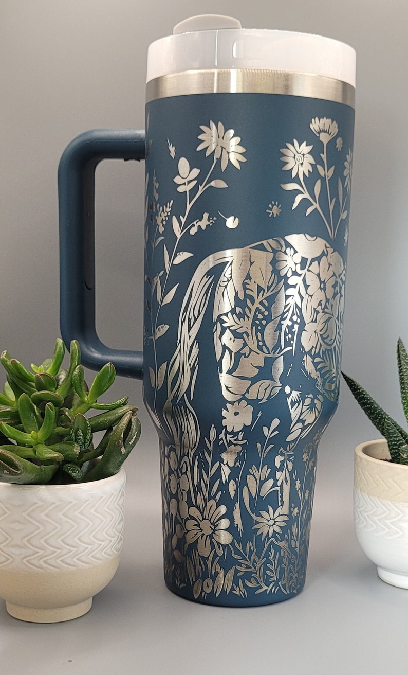 May include: A blue stainless steel travel mug with a silver floral design featuring an elephant. The mug has a lid and a handle.
