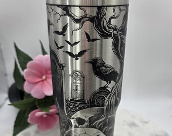 May include: Stainless steel travel tumbler with a black and silver engraved design. The design features a graveyard scene with a raven, bats, tombstones, and skulls. The tumbler has a black lid and a silver rim.
