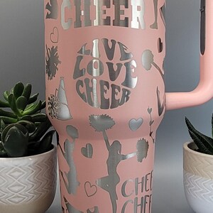 May include: A pink stainless steel tumbler with a silver design featuring cheerleaders, pom-poms, and the text "CHEER LIVE LOVE CHEER".